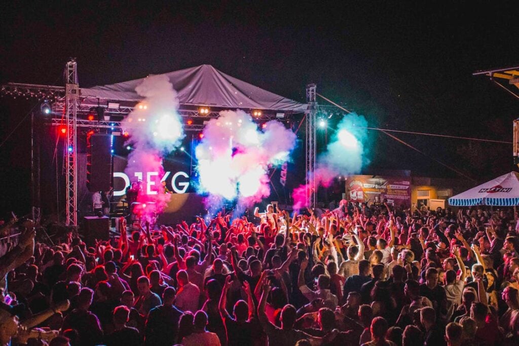 A vibrant concert scene with colorful lights and smoke above a large, energetic crowd facing a lit-up stage at night.