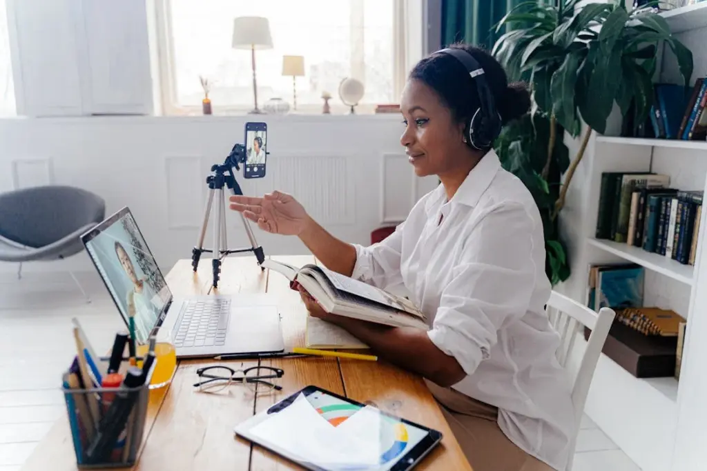 A person wearing headphones sits at a desk with a laptop, smartphone on a tripod, tablet, and book, engaged in a video call. The background includes shelves with books and a large plant.