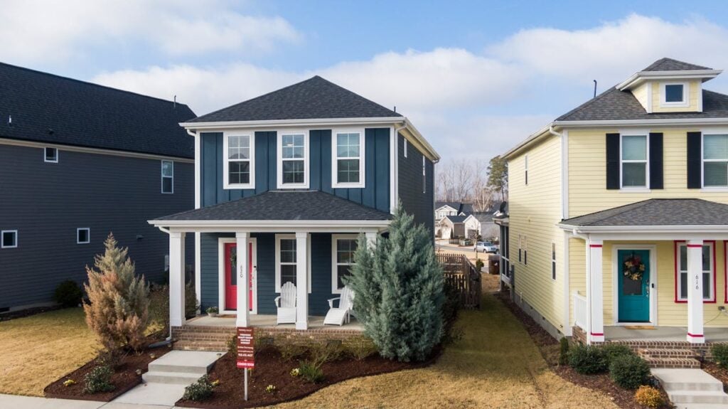 A two-story blue house with a front porch, flanked by neighboring homes. A for-sale sign is displayed in the front yard.