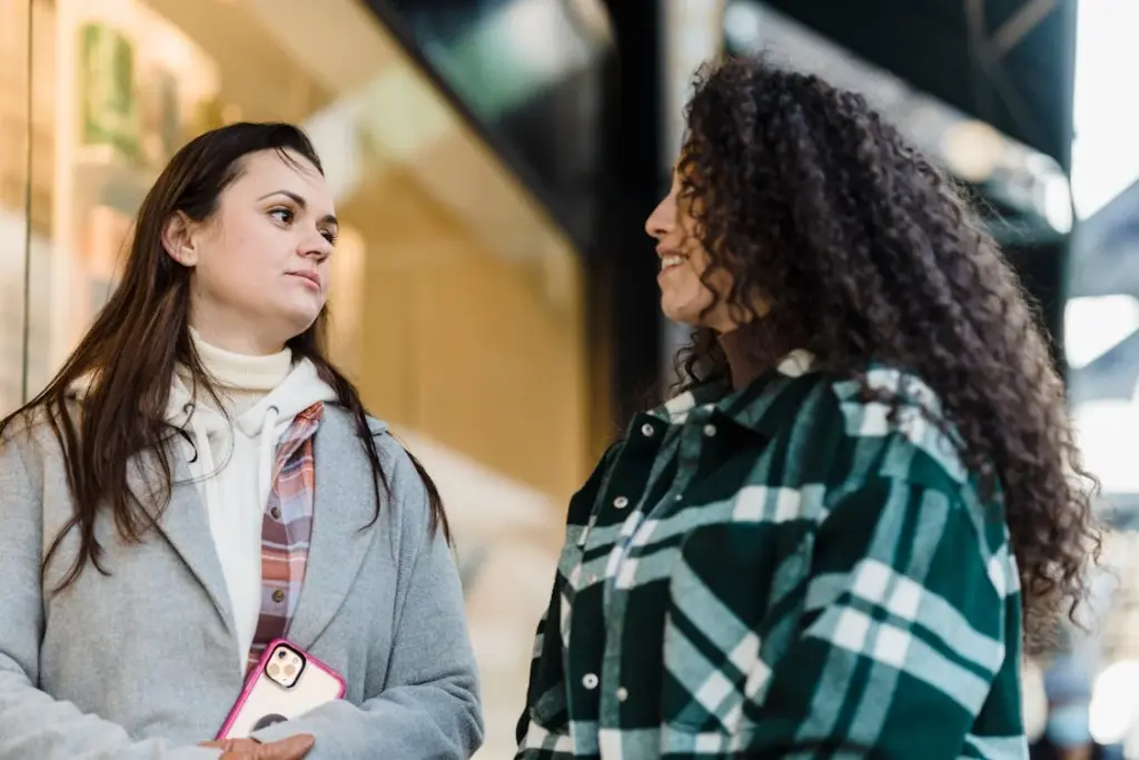 Two women are standing and talking outdoors. One woman holds a smartphone, while the other smiles at her. Both are dressed in warm clothing.