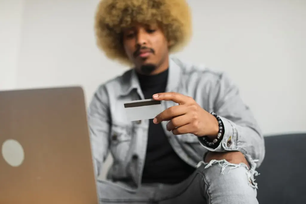 A person with an afro hairstyle uses a laptop while holding a credit card in their hand. They are dressed in a denim jacket and jeans.