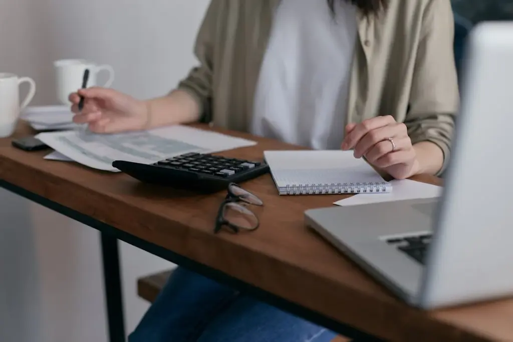 A person sits at a desk with a notebook, papers, calculator, eyeglasses, and a laptop, holding a pen. Two coffee cups are in the background.