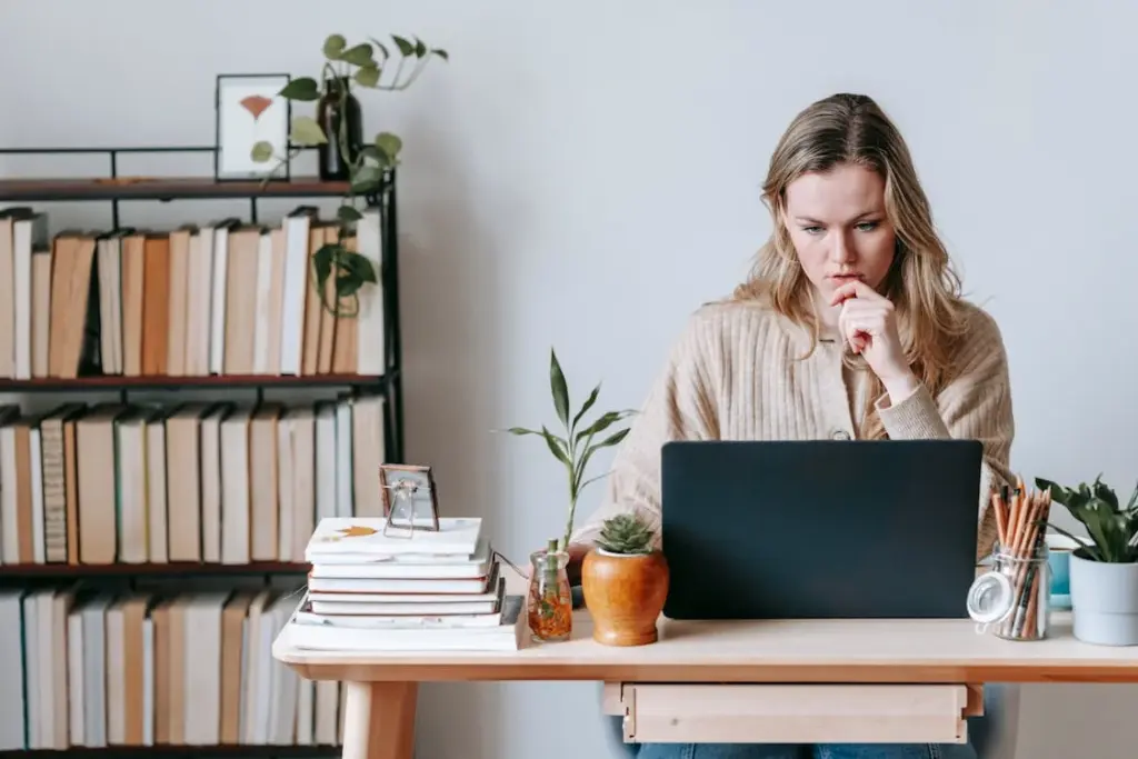 A woman sits at a desk with her hand on her chin, working on a laptop. The desk has books, plants, and stationery. A bookshelf filled with books is in the background.