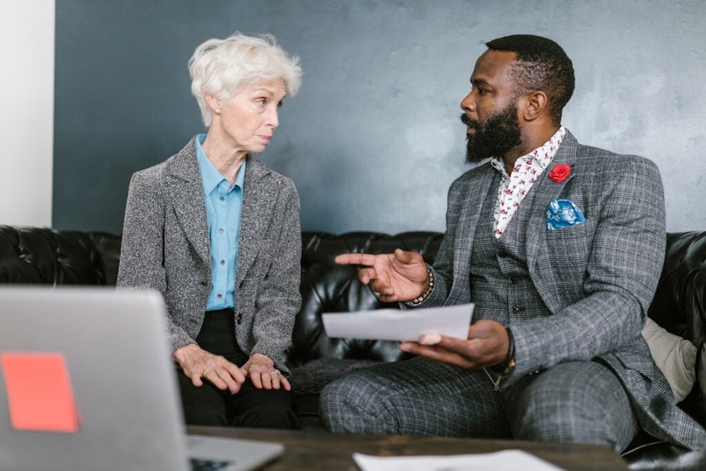 Two people in business attire sit on a sofa, engaged in a conversation. One holds a tablet.