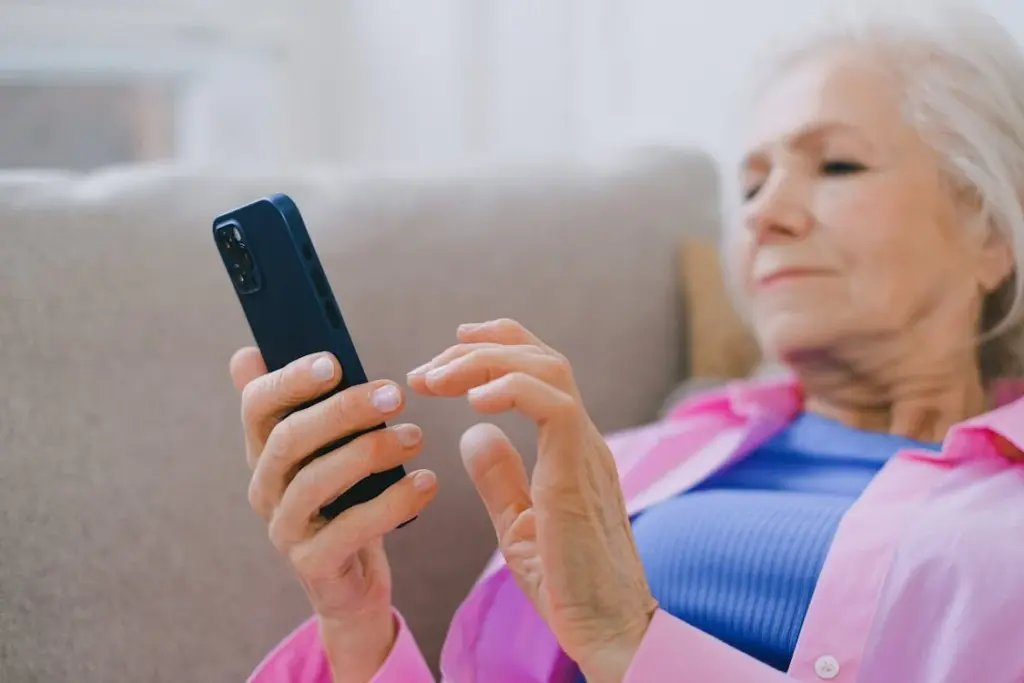 Elderly woman in a pink shirt using a smartphone while reclining on a beige sofa.