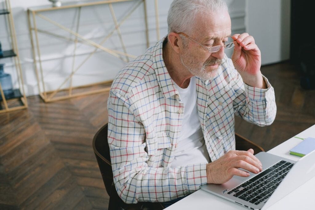 An older man with gray hair and glasses is using a laptop at a desk. He is wearing a plaid shirt and appears focused on the screen, perhaps researching why companies do not hire over 50.