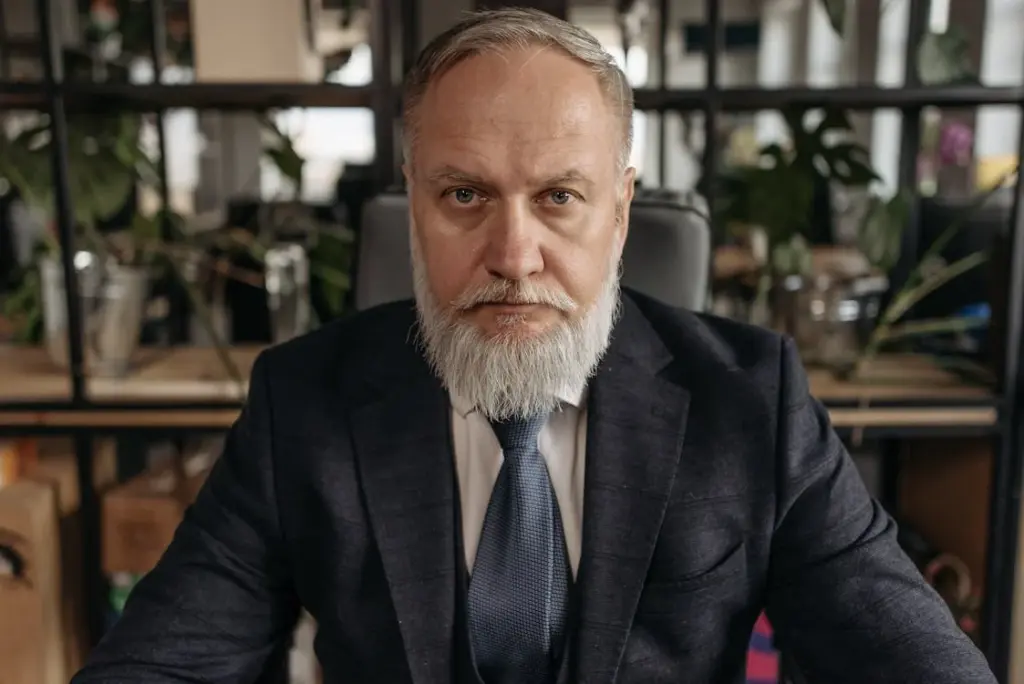 A man with a gray beard and mustache, wearing a dark suit and tie, sits in an office with shelves and plants in the background, pondering why companies do not hire over 50.