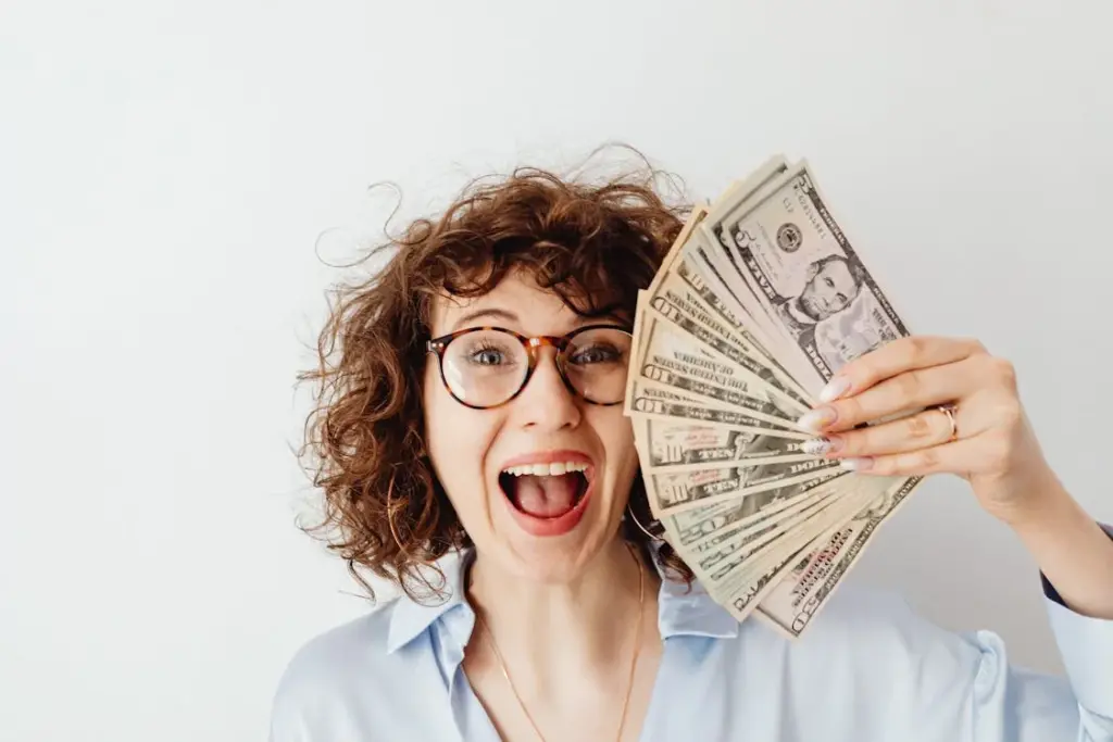 A person with curly hair and glasses smiles enthusiastically while holding an assortment of U.S. dollar bills fanned out in one hand, highlighting the irony of things you're paying for but can get for free.