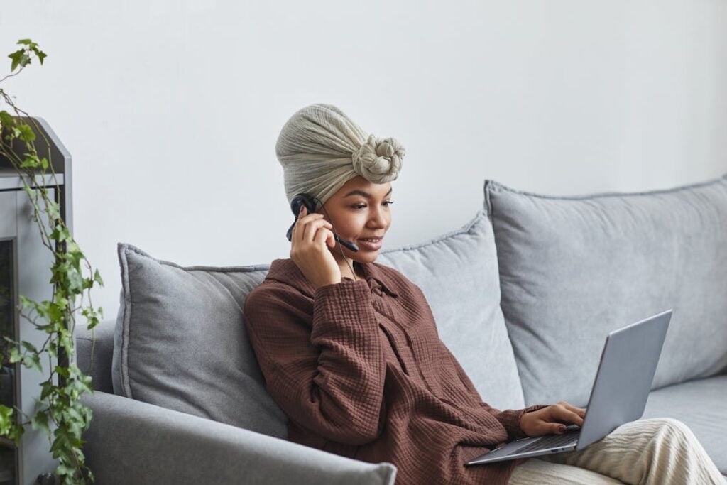 A person sits on a couch, talking on the phone and using a laptop. They wear a brown shirt and a light-colored headwrap. A plant is visible beside the couch.