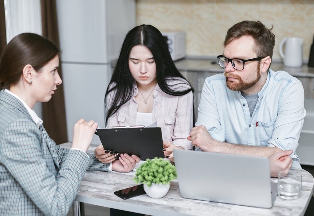 Three people sitting at a table with a laptop and clipboard, discussing something serious.