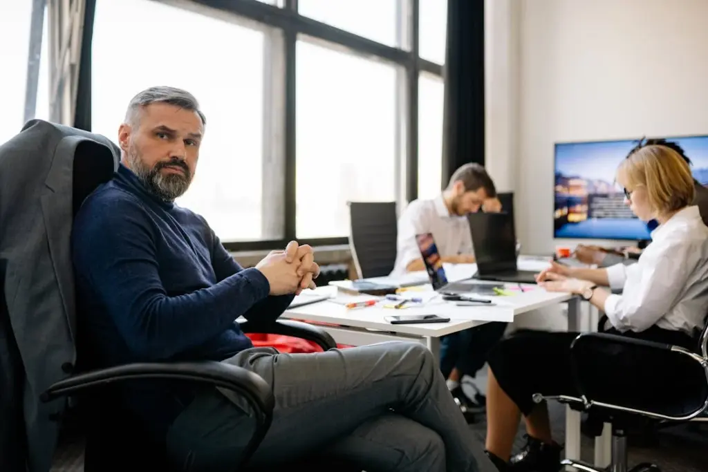 A bearded man sits at a desk in an office, with two colleagues working at computers in the background.