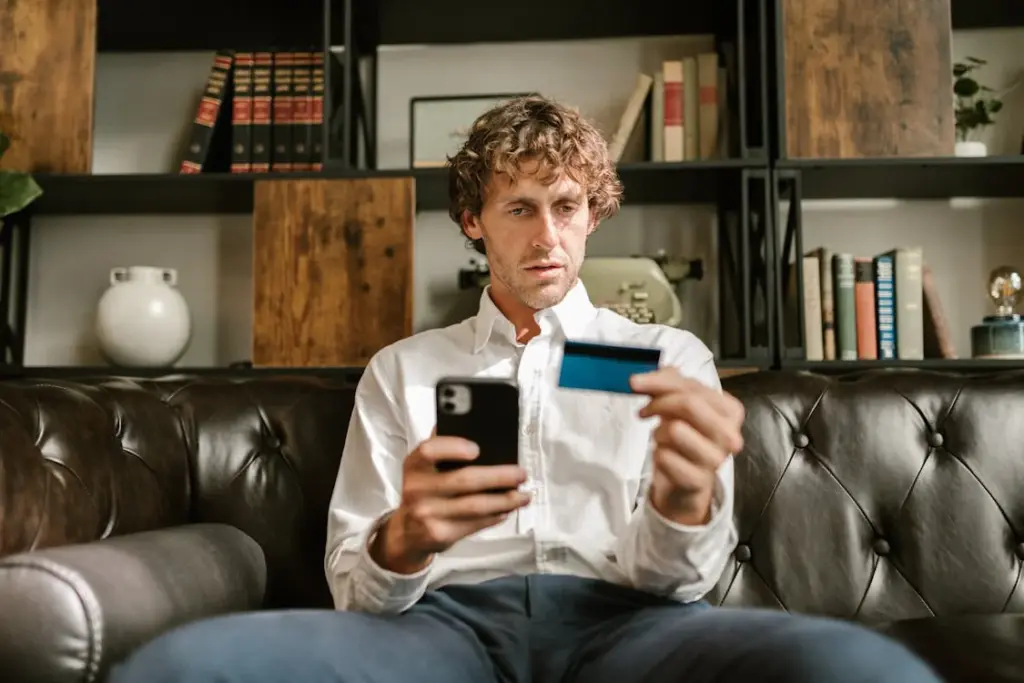 A man in a white shirt sits on a leather couch holding a smartphone in one hand and a credit card in the other, with bookshelves in the background.