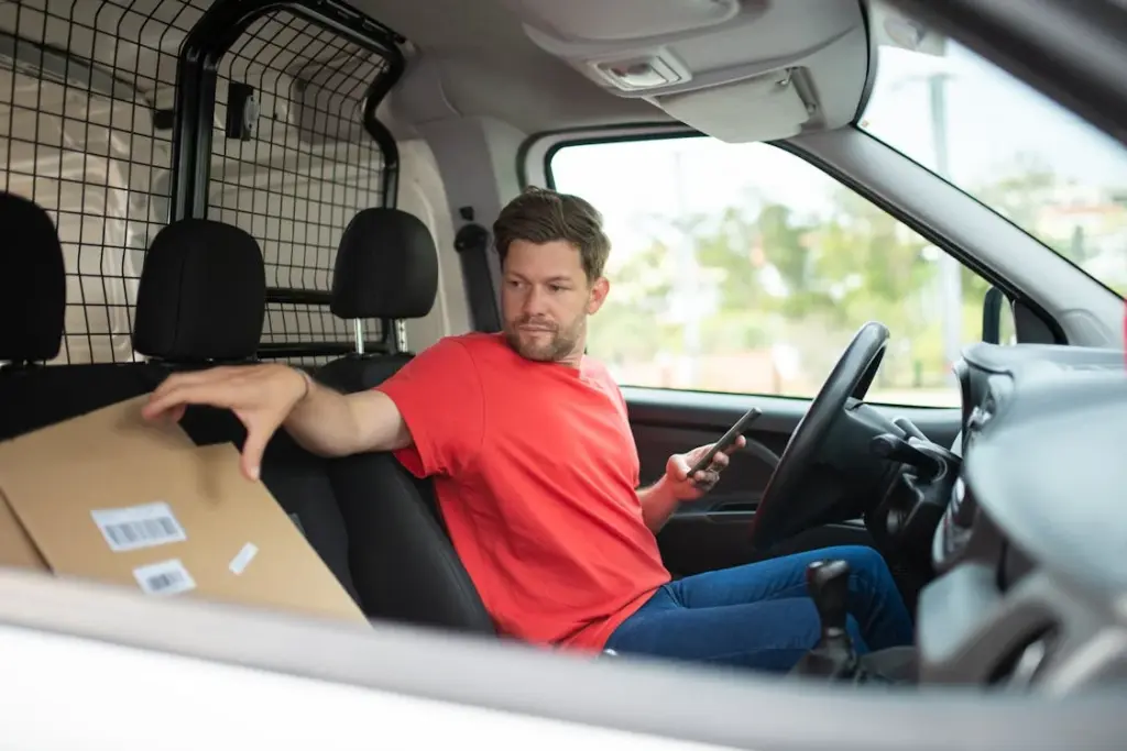 A man in a red shirt is sitting in the driver's seat of a vehicle. He is holding a smartphone in one hand and reaching for a package on the passenger seat with the other.