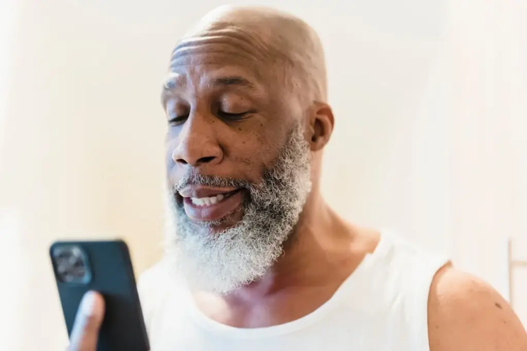 A man with a beard looks at his phone, wearing a white tank top.