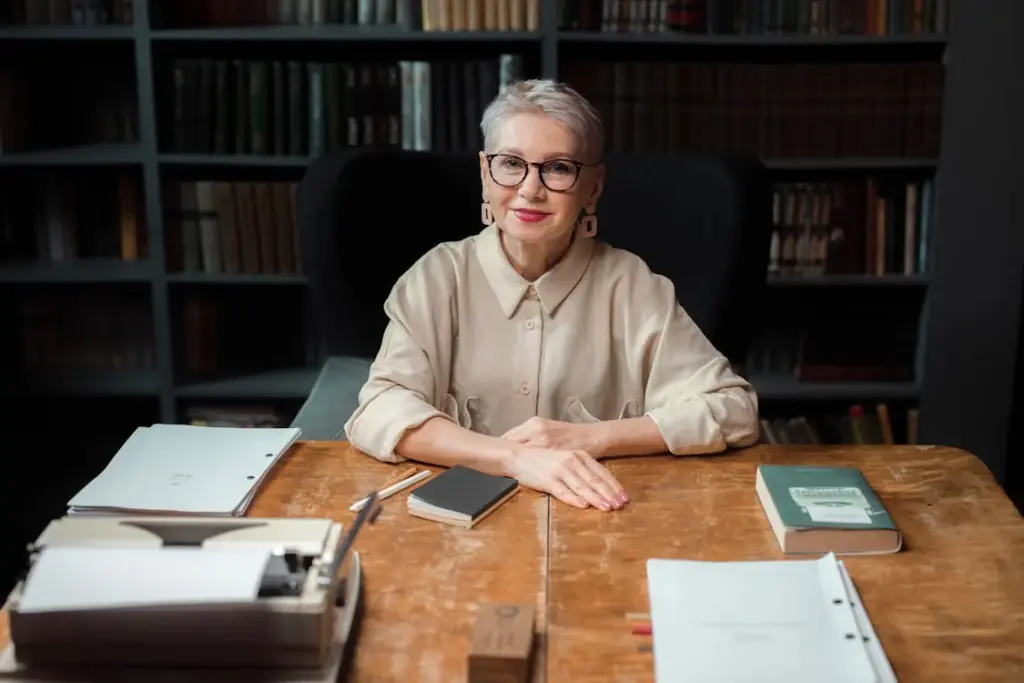 An older woman with short gray hair, wearing glasses and a beige shirt, sits at a wooden desk in an office with shelves of books behind her, accompanied by documents, a typewriter, and a notebook.