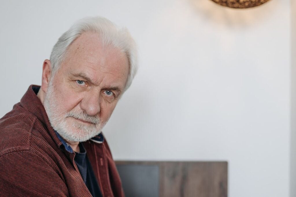 An elderly man with white hair and beard wearing a maroon shirt looks into the camera with a neutral expression in a well-lit room.