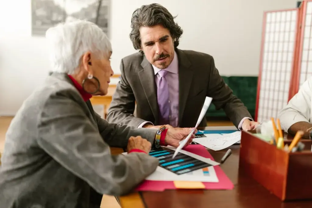 An elderly woman and a middle-aged man in a suit discuss documents at a table in an office setting.