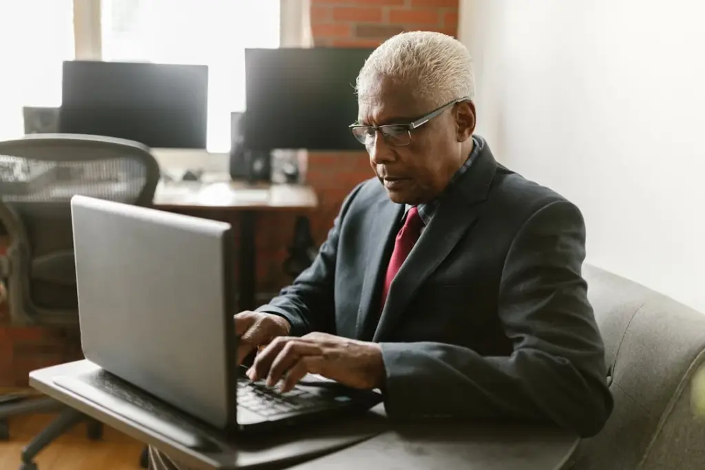 An elderly man wearing a suit and tie types on a laptop in a modern office space. Two computer monitors are visible in the background.