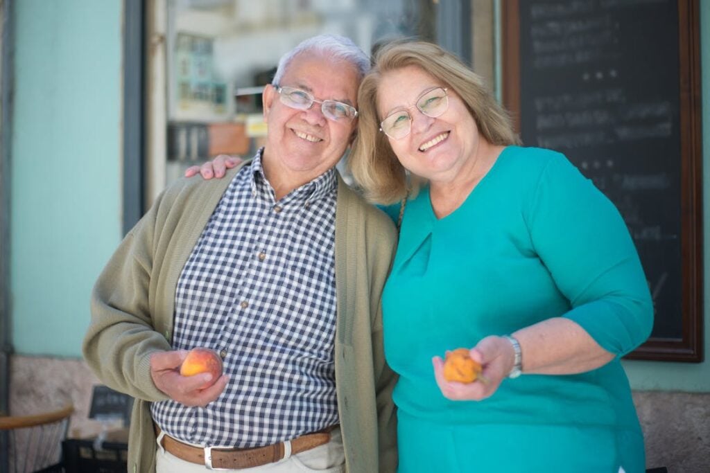 An elderly man and woman, both smiling, stand together outside a building while each holding a peach in their hands.