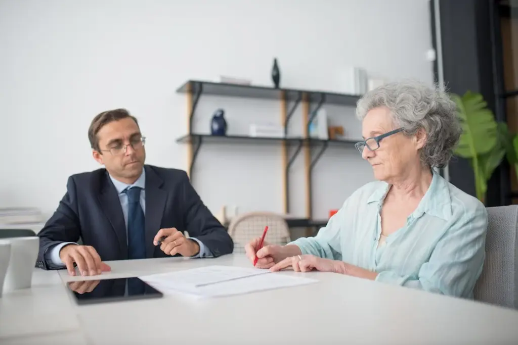 An older woman signs a document with a red pen while a man in a suit observes, seated at a table in an office setting.