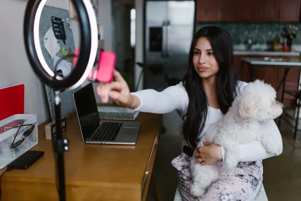 Woman holding a white dog sits at a desk, adjusting a smartphone on a ring light stand. A laptop is open nearby in a home office setting.