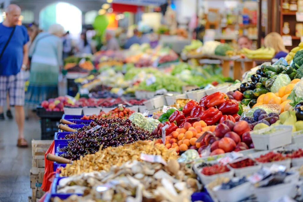 A bustling market scene with displays of fresh fruits and vegetables, including peppers, peaches, cherries, and greens, and people browsing in the background.