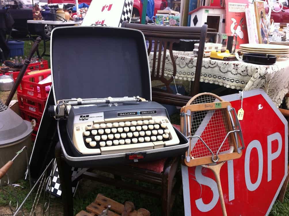 A vintage typewriter in an open case is displayed at a flea market, surrounded by various items like a wooden tennis racket, a red stop sign, and boxes.