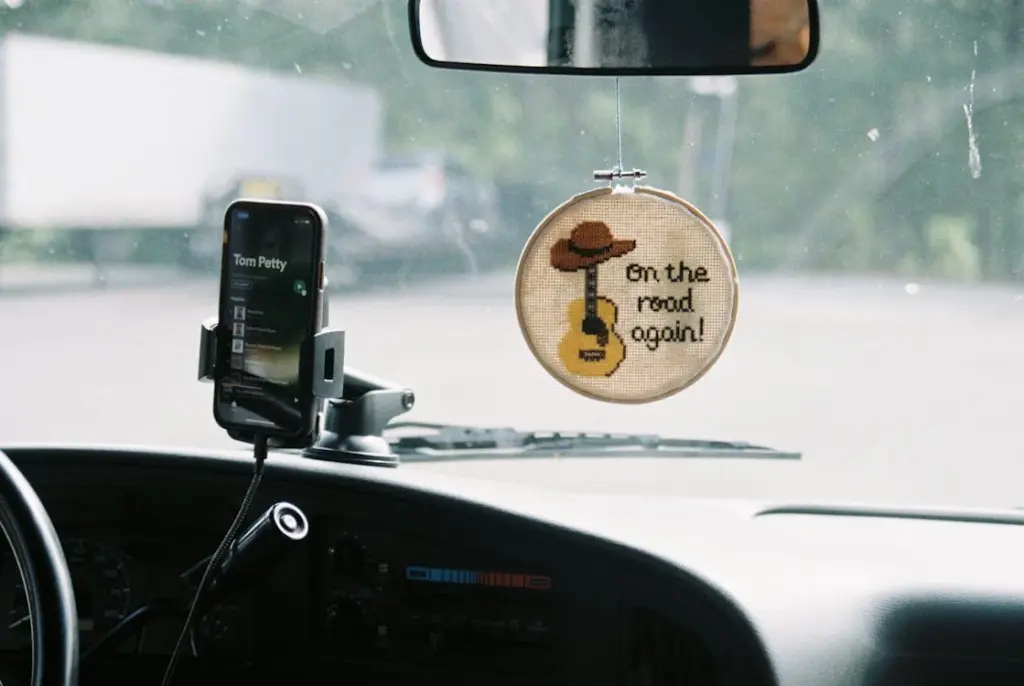 Car dashboard view with a mounted phone showing Tom Petty on the screen. A cross-stitch hoop displays 