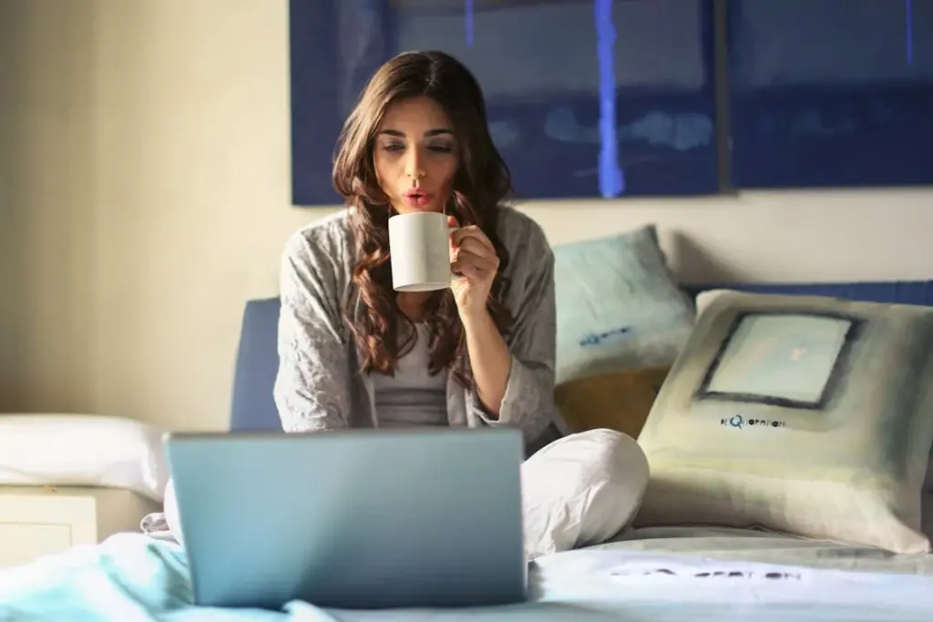 Woman sits on a bed with pillows, drinking from a white mug, and looks at a laptop screen.