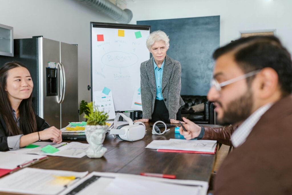 An elderly woman stands by a table with three younger adults seated, engaged in a discussion. A whiteboard with notes, a refrigerator, and office supplies are visible in the background.