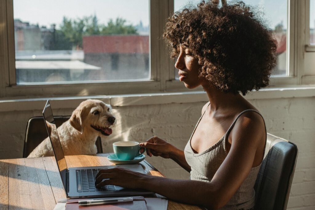 A person is sitting at a table, working on a laptop with a coffee cup in hand. A dog is beside the person, looking outside through a window.