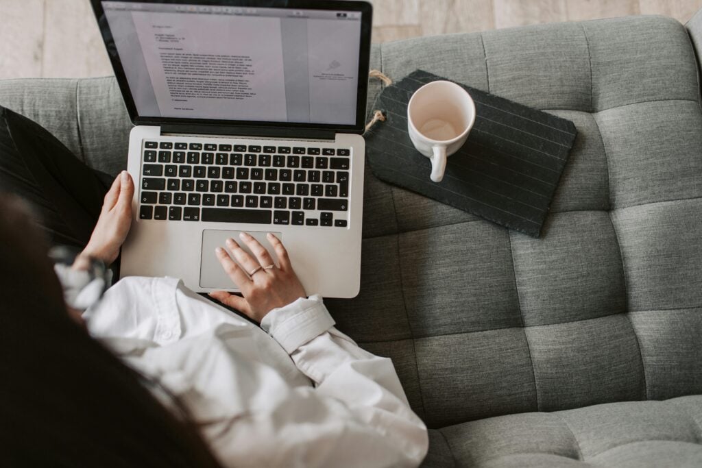 A person is sitting on a gray couch using a laptop with a document open. A mug and a notepad are placed beside the laptop.