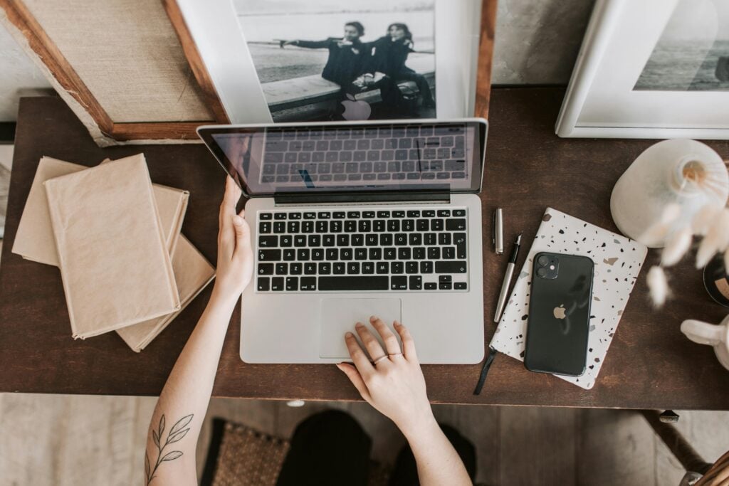 Person using a laptop on a wooden desk with books, a notebook, an iPhone, and a framed photo nearby.