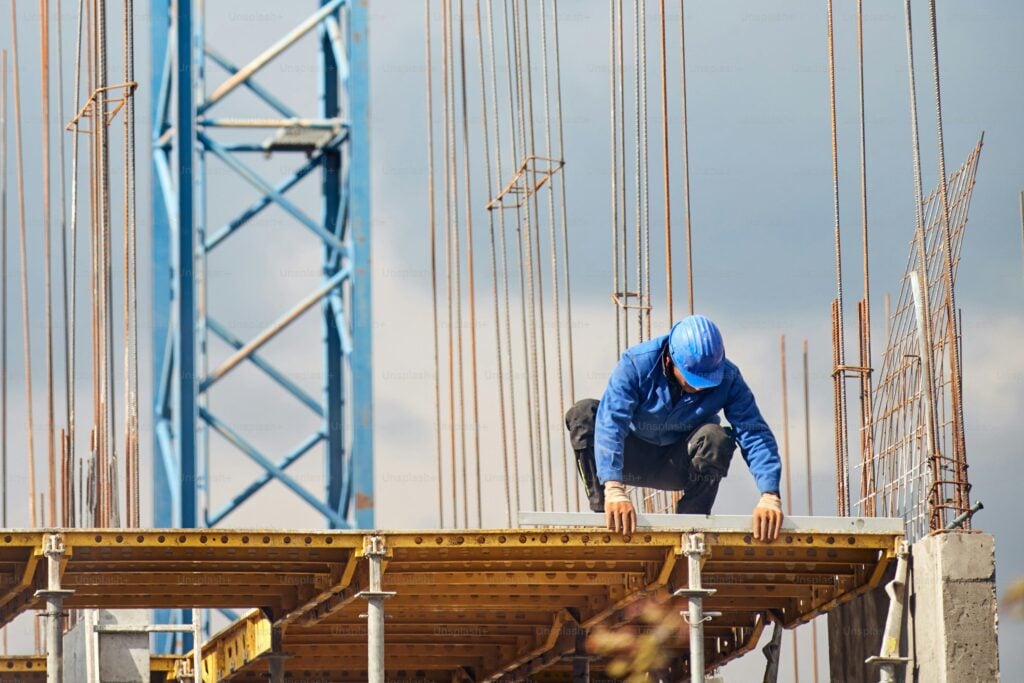 A construction worker in a blue helmet crouches on a scaffolding platform with steel rods and a crane in the background.