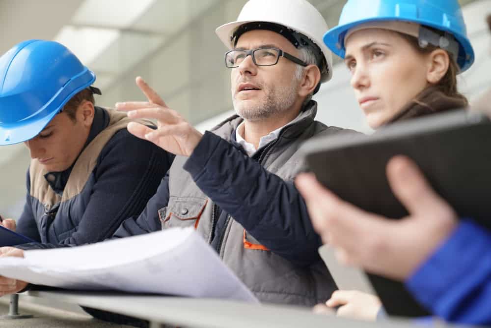 Three construction professionals in hard hats review blueprints and discuss the project at a construction site.