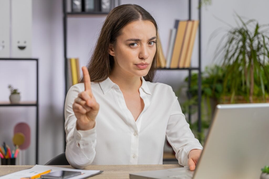 A person with long hair, wearing a white shirt, sitting at a desk with a laptop, holds up one finger in a gesture indicating 