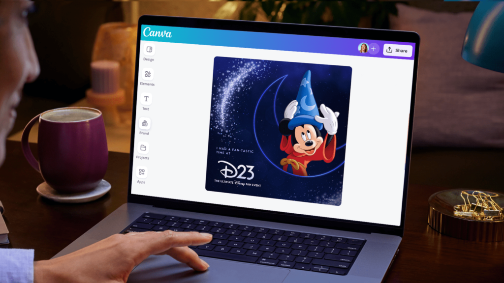 Person using a laptop displaying a Canva design of Disney's Mickey Mouse in a wizard hat, with a coffee mug and a lamp nearby.