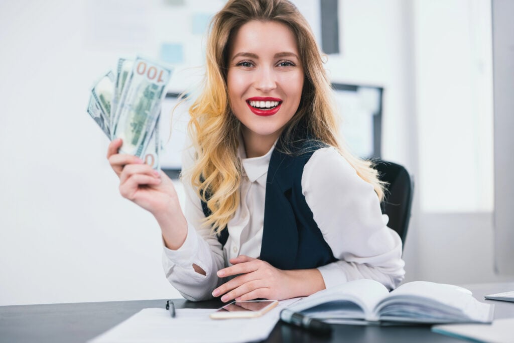 A woman sitting at a desk holds multiple hundred-dollar bills in her hand, smiling. An open book and pen are visible in front of her.