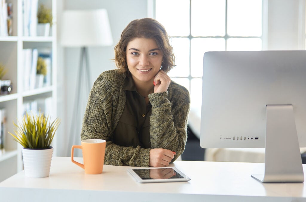 A person with short hair smiles, leaning on a desk with a computer, tablet, and orange mug, in a bright room with a window.