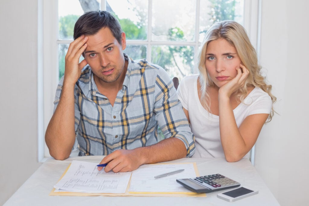 A man and woman sit at a table with financial documents and a calculator, both looking stressed.