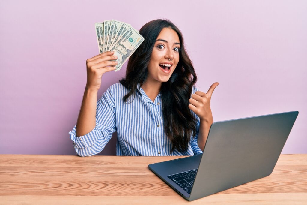 A woman holds fan of dollar bills in one hand and gives a thumbs up with the other while sitting at a table with an open laptop.