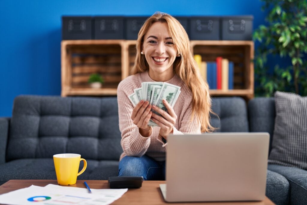 A person sitting on a couch holds a stack of dollar bills, with a laptop and papers on a table.