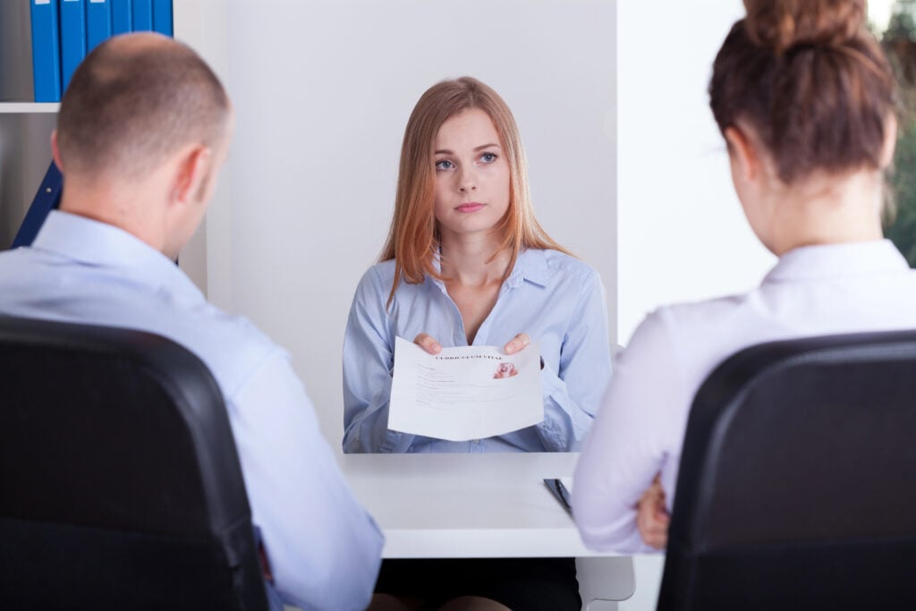 A woman holding a resume sits across a table from two interviewers in an office setting.