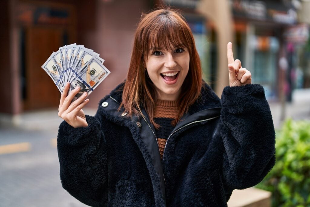 A woman with brown hair is smiling and holding a fan of US $100 bills in one hand while pointing upwards with the other hand. She is wearing a black coat and standing outdoors.