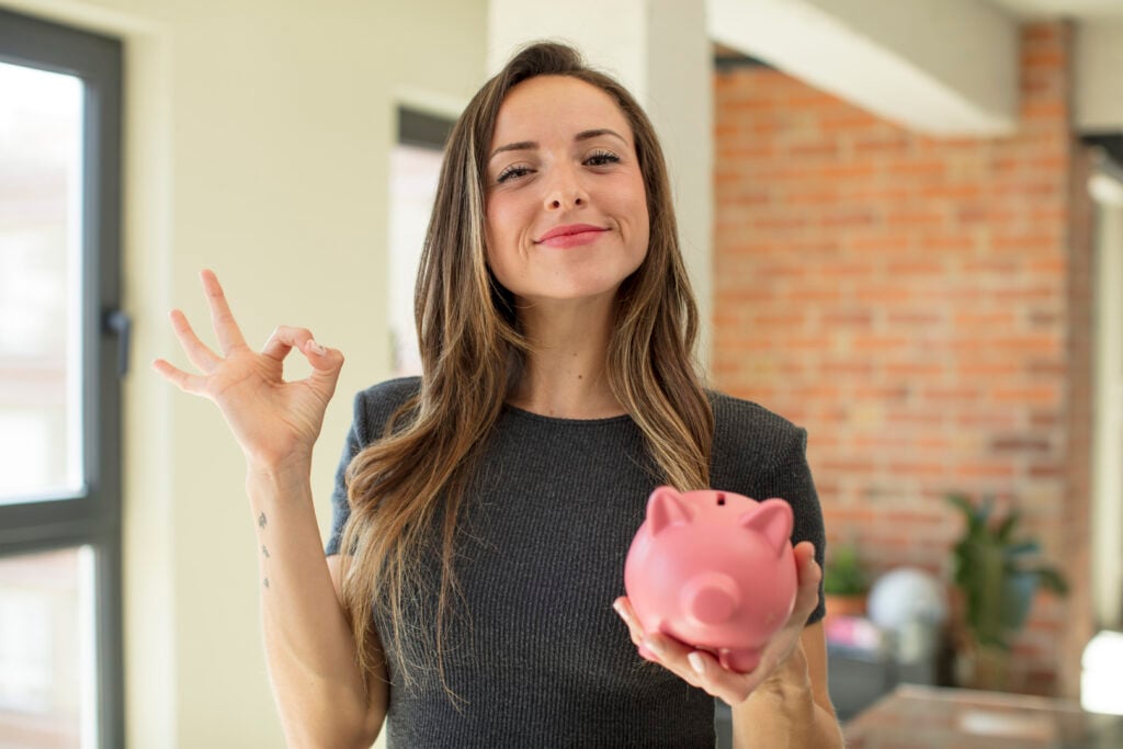 A woman smiles while holding a pink piggy bank in one hand and making an 