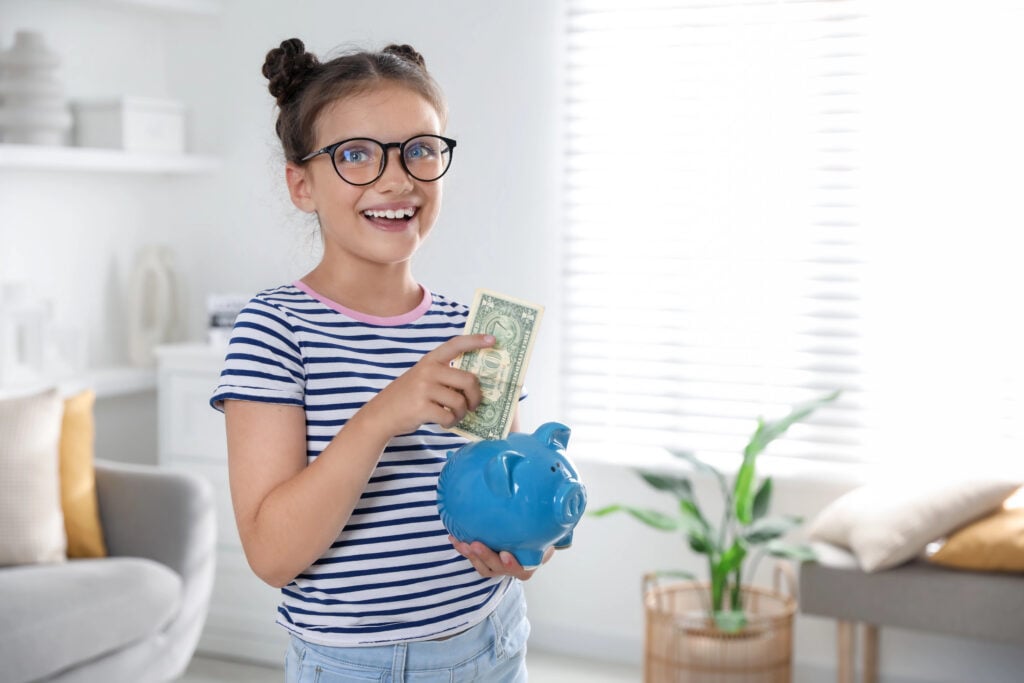 Girl with glasses placing a dollar bill into a blue piggy bank, smiling in a brightly lit living room.