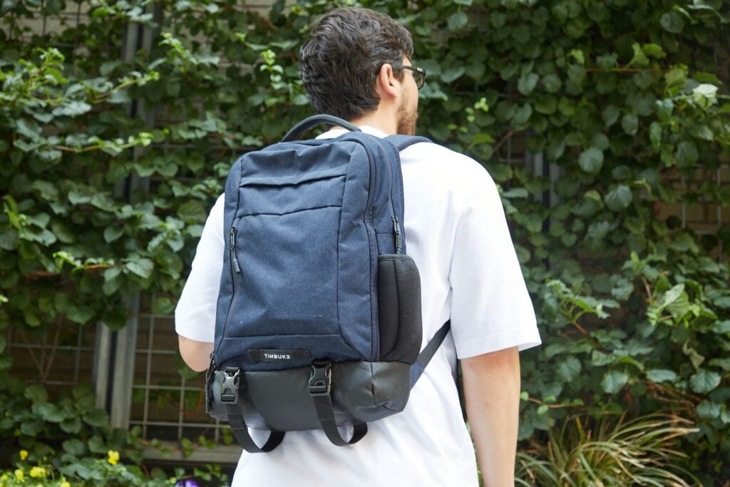 Person wearing glasses and a white shirt, carrying a blue backpack, stands in front of a leafy background.