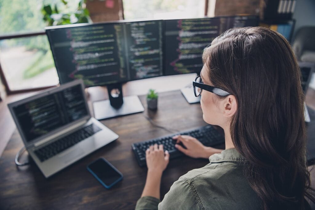 Person at a desk using a desktop computer with multiple screens displaying code. A laptop and a smartphone are also on the desk.