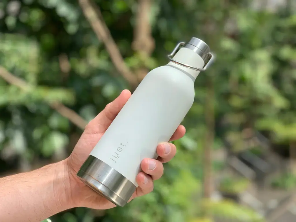 A hand holding a white insulated water bottle outdoors with blurred green foliage in the background.