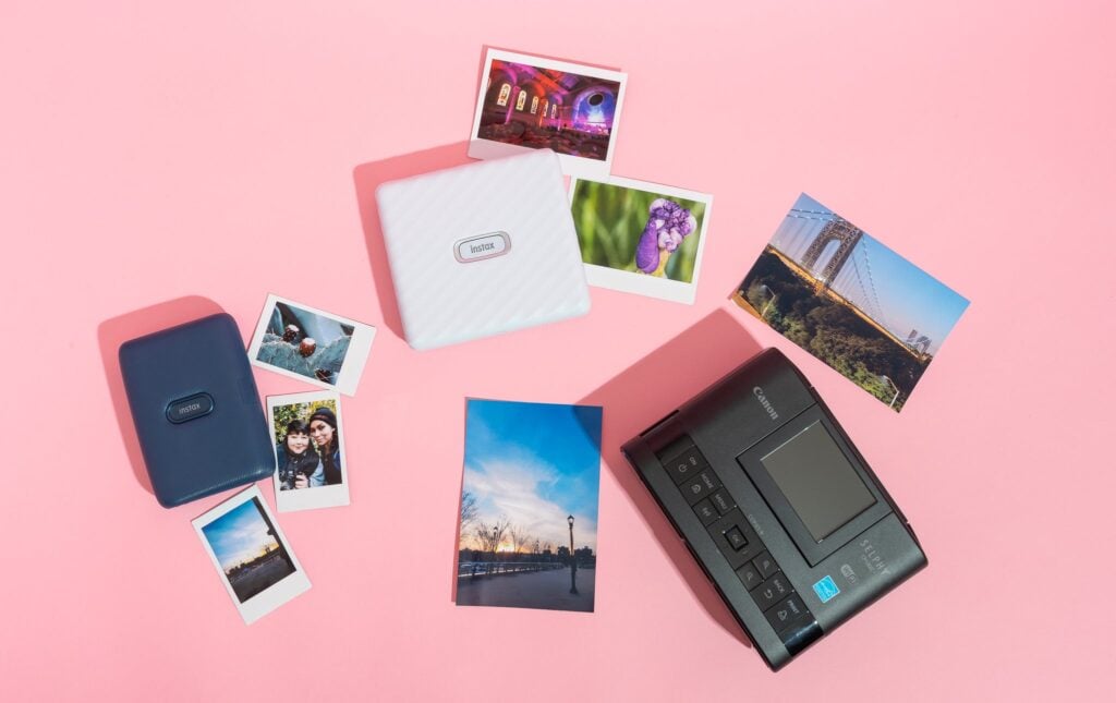 A flat lay of portable photo printers, printed photos, and a dark gray device on a pink background.